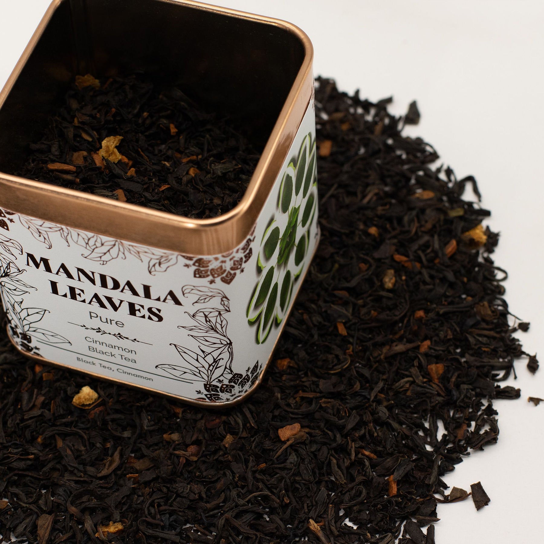 Tea tin labeled 'Mandala Leaves' surrounded by black tea leaves on a light background