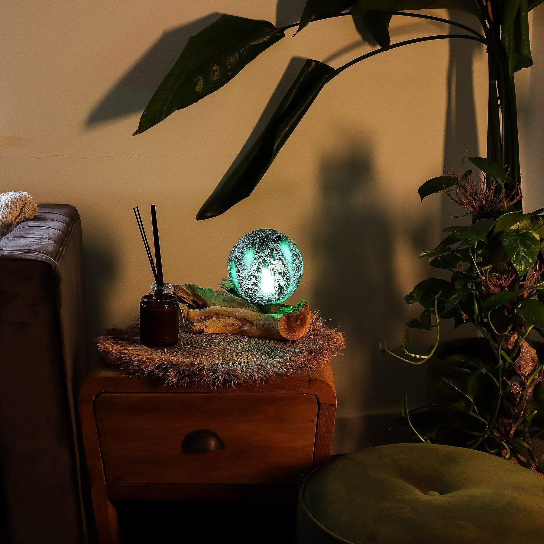 Emerald crackle glass sphere glowing atop unique twisted driftwood base on dark wooden side table with books and plants at night