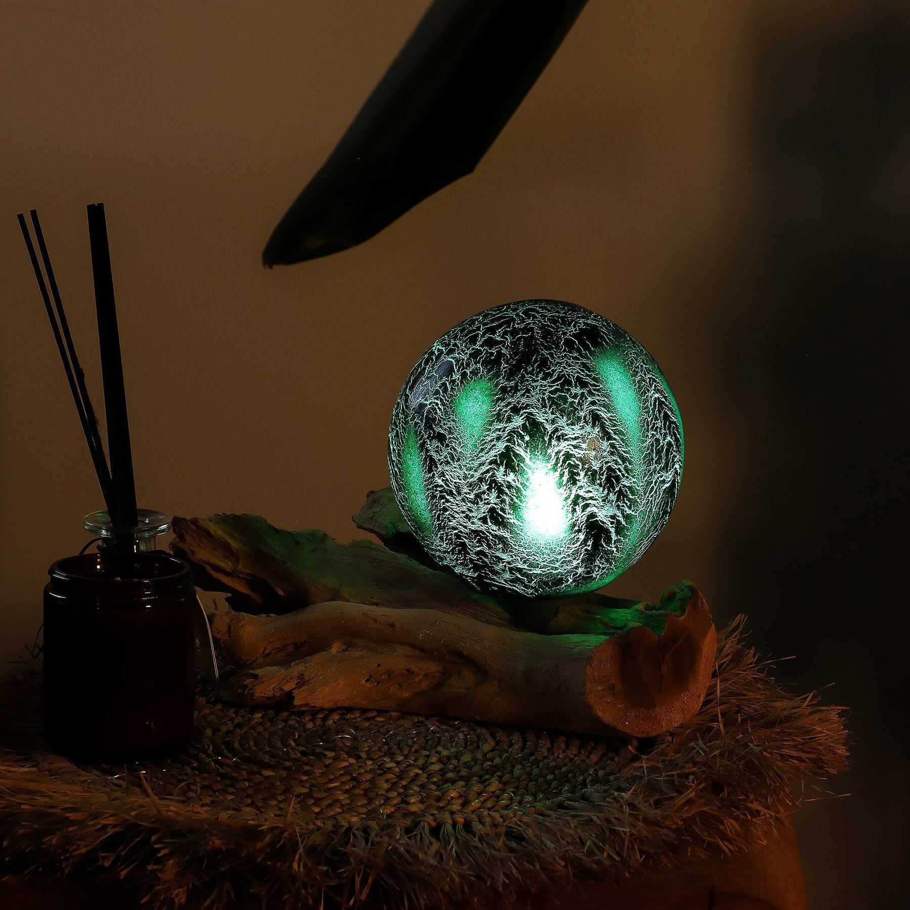 Emerald crackle glass sphere glowing atop unique twisted driftwood base on dark wooden side table with books and plants at night