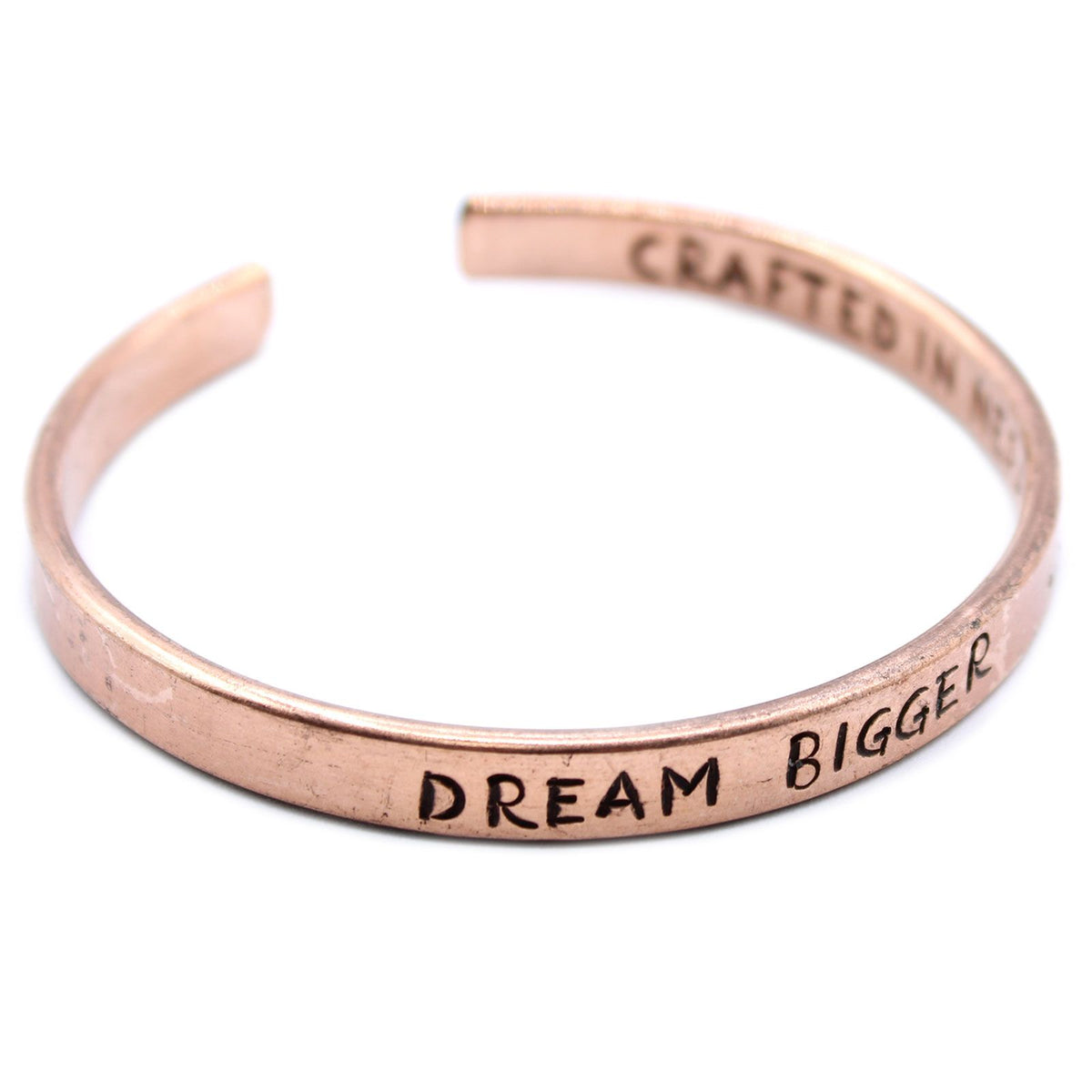 Open copper mantra cuff bracelet engraved “DREAM BIGGER” on wrist, surrounded by Himalayan prayer flags and mountains in soft sunrise light