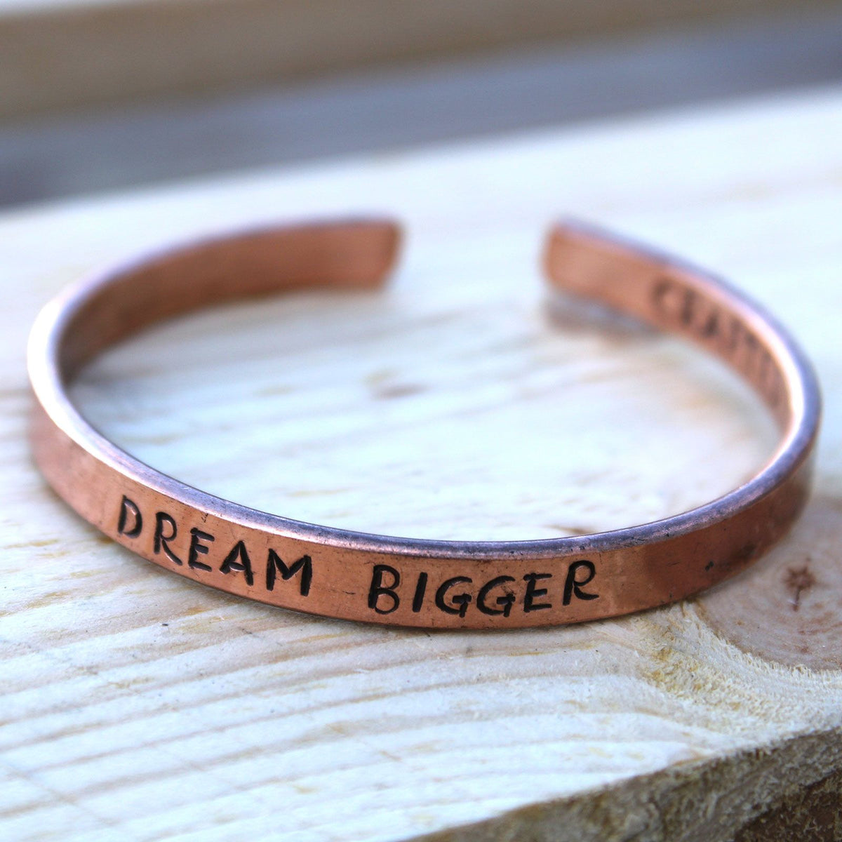 Open copper mantra cuff bracelet engraved “DREAM BIGGER” on wrist, surrounded by Himalayan prayer flags and mountains in soft sunrise light