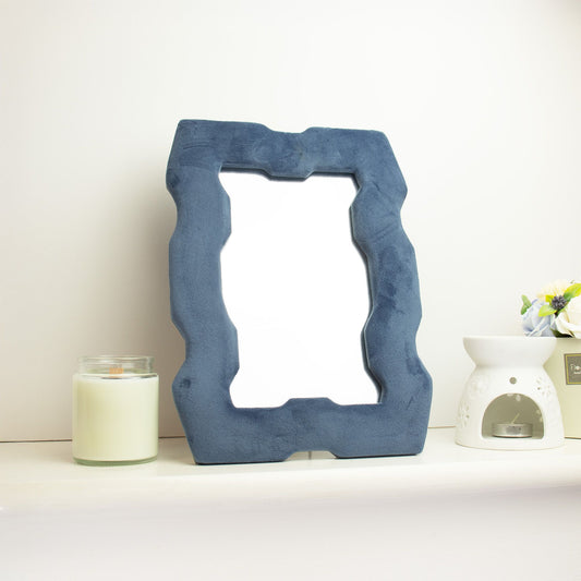 Soft squiggly mirror in rich royal blue velvet chunky frame with wavy sculpted edges, freestanding on white dressing table with soft natural light reflecting in glass