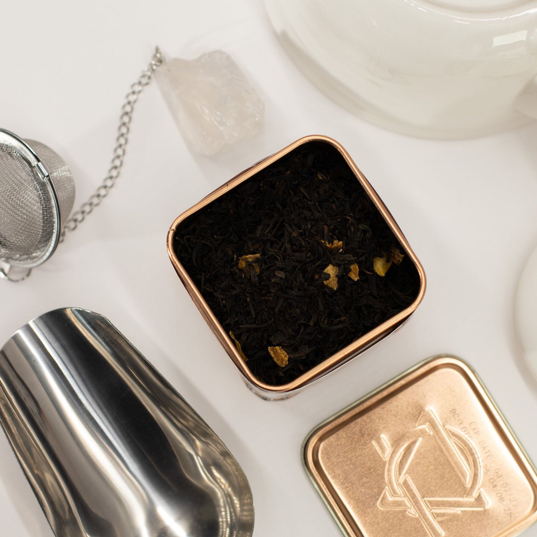 Tea leaves in a small square container with a silver tea strainer and white teacup in the background.