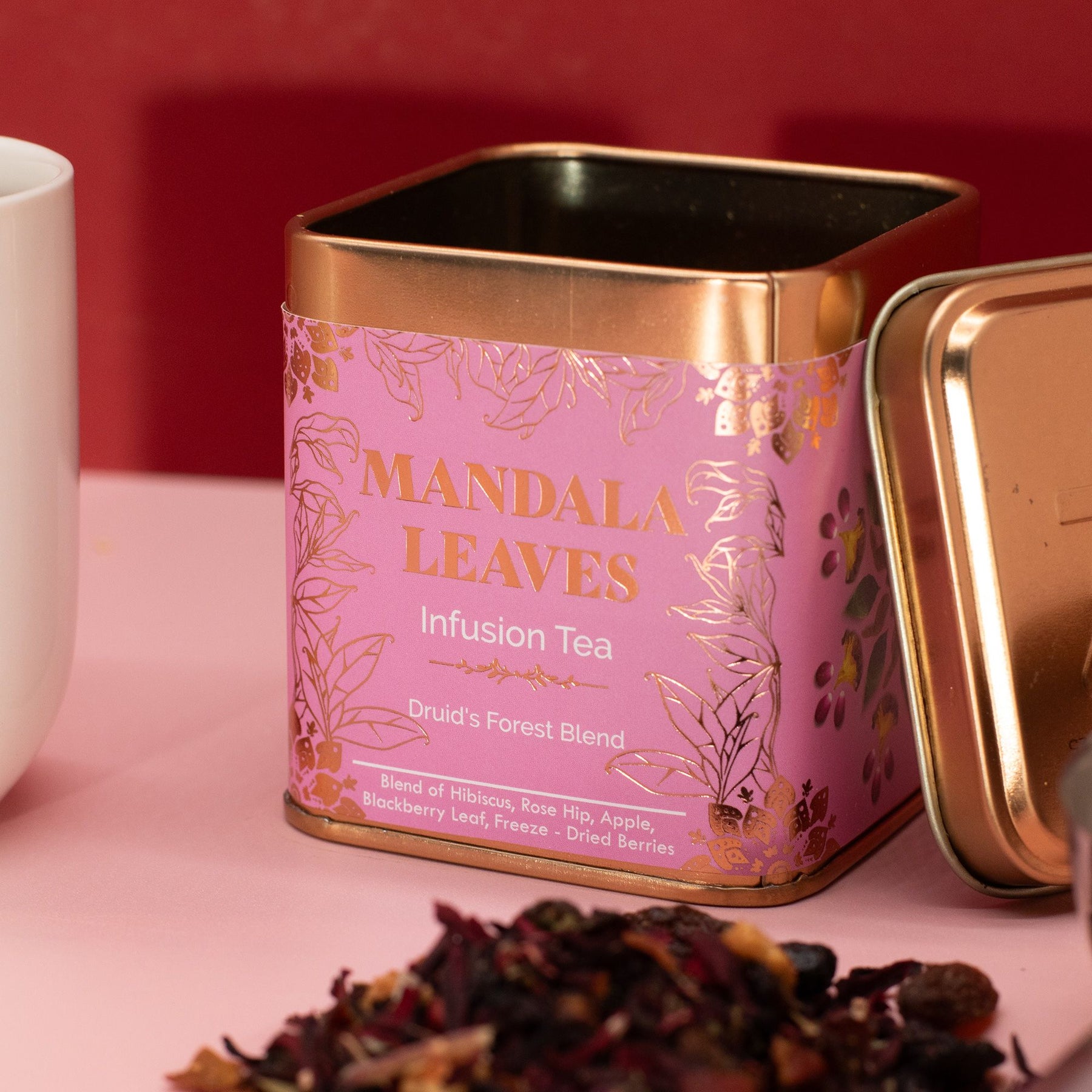 Pink tea canister labeled 'Mandala Leaves' with a white mug and tea leaves on a pink surface.