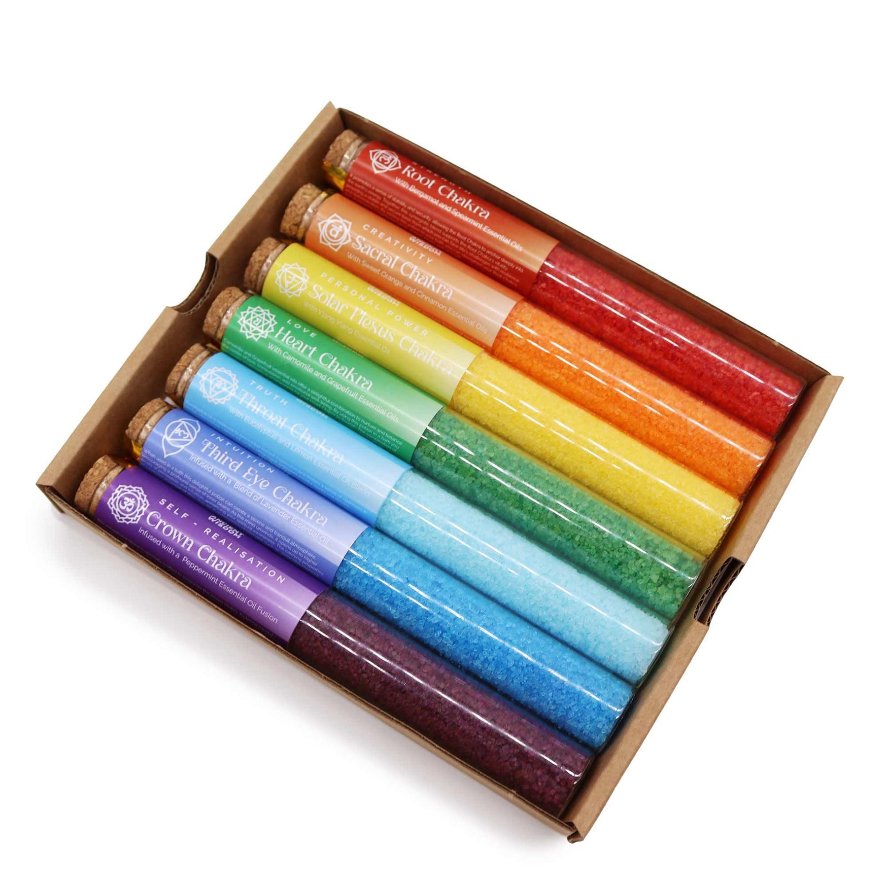 Rainbow row of 7 coloured chakra bath salt vials with essential oil droppers and crystals, arranged on white marble with candles and rose petals