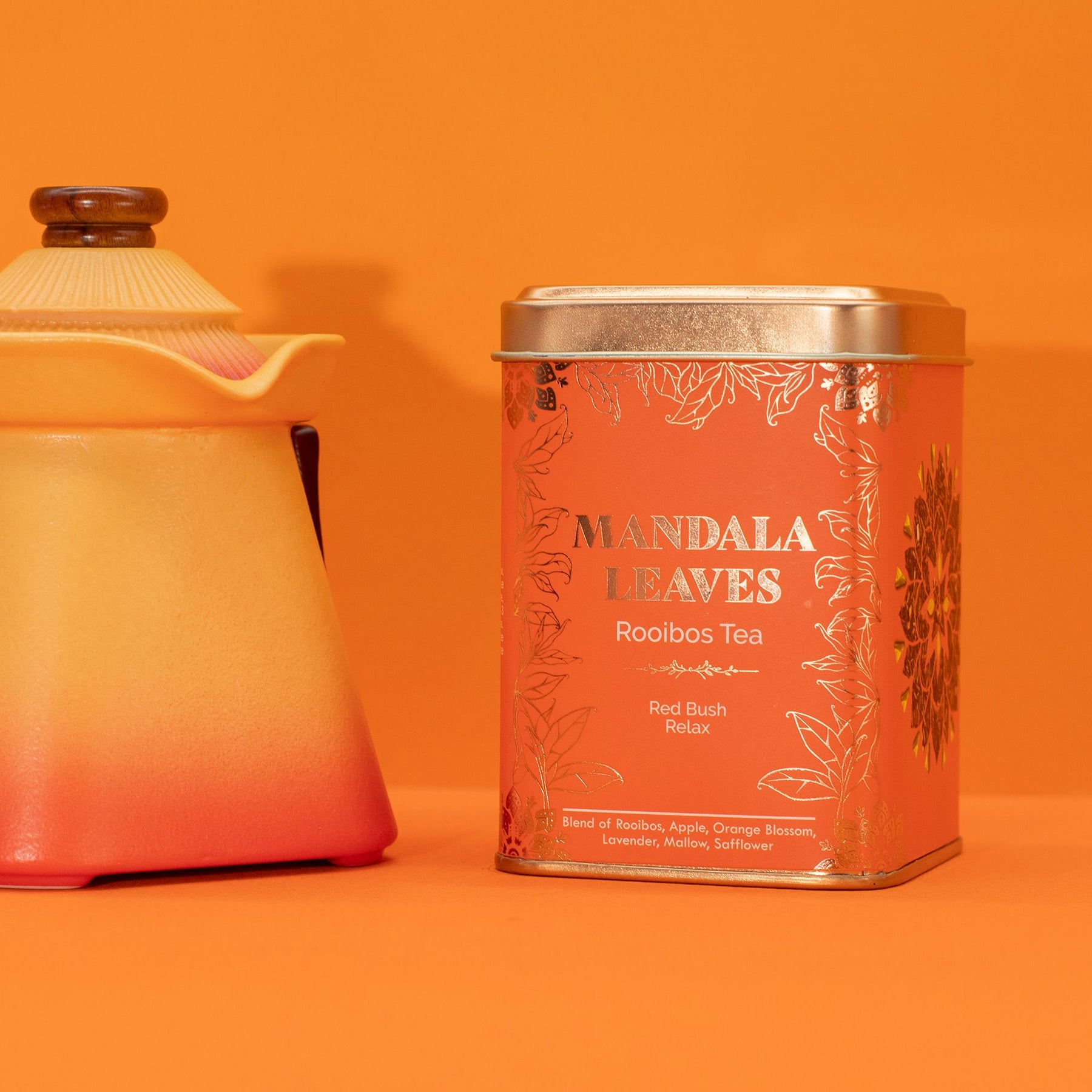 Tea canister labeled 'Mandala Leaves Rooibos Tea' next to a ceramic pitcher on an orange background