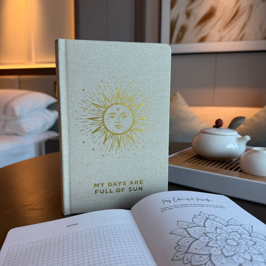 Notebook with sun design and text 'My days are full of sun' on a table with a teapot and open book.