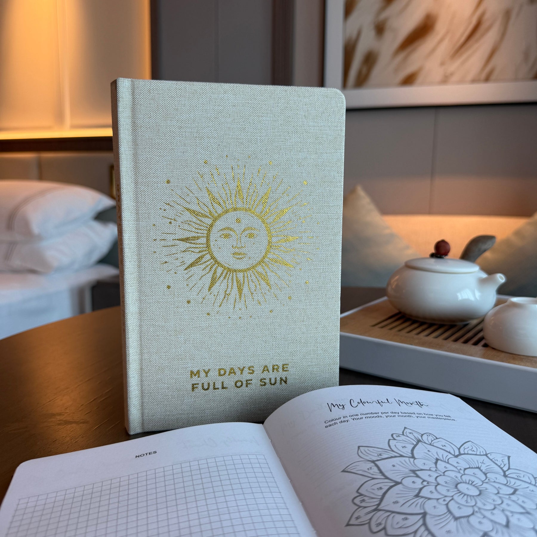 Notebook with sun design and text 'My days are full of sun' on a table with a teapot and open book.