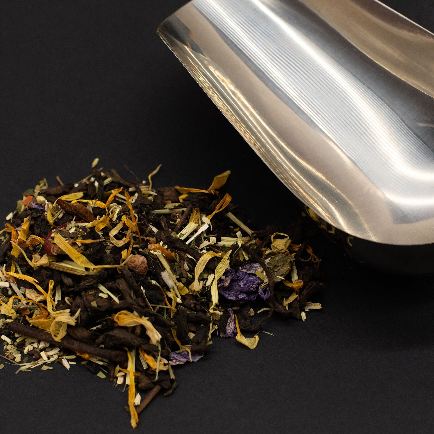 Metallic tea scoop over a pile of dry tea leaves on a black background