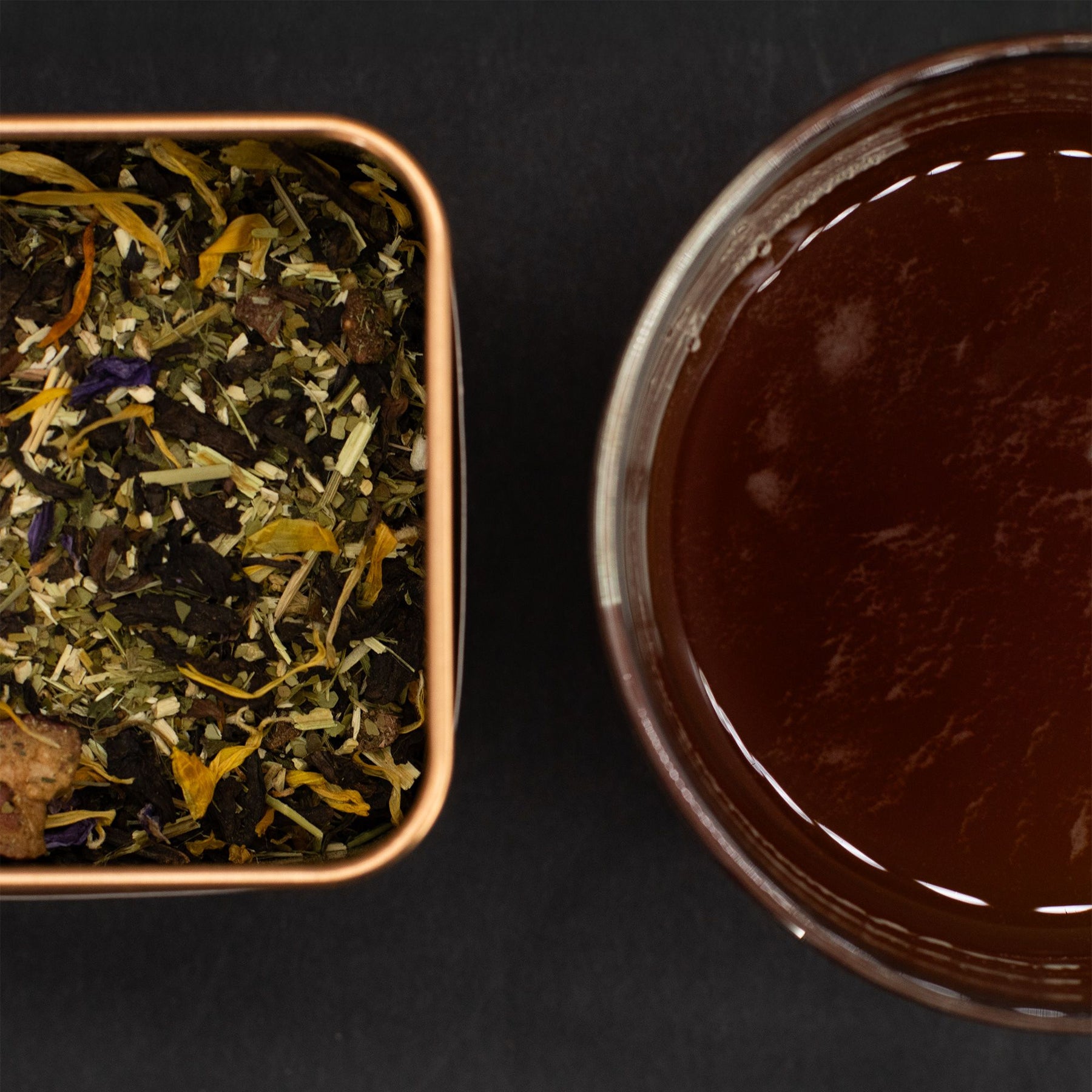 Tea leaves in a container next to a cup of brewed tea on a dark surface