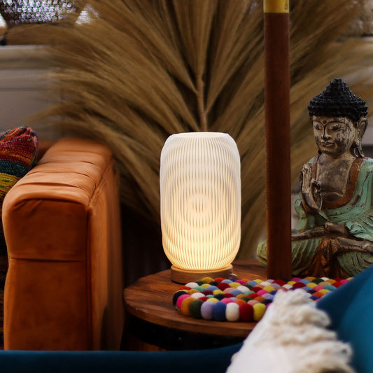 Decorative Modern Twisted New Wave USB lamp with textured honeycomb surface and sleek twisted silhouette, warm glow on 3rd brightness setting, placed on a wooden base on a table with a Buddha statue and colorful items in a cozy room setting.
