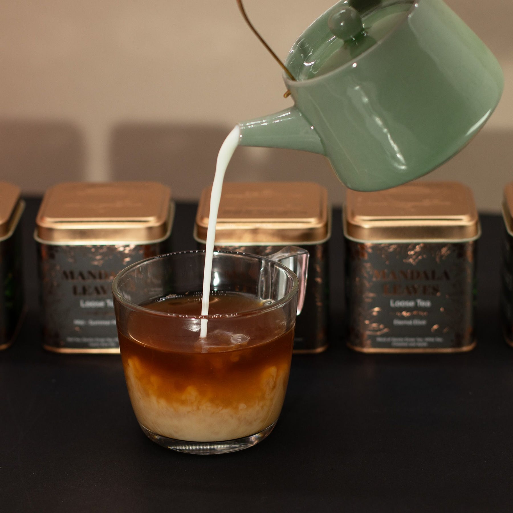 Green teapot pouring milk into a glass of iced coffee with tea tins in the background.