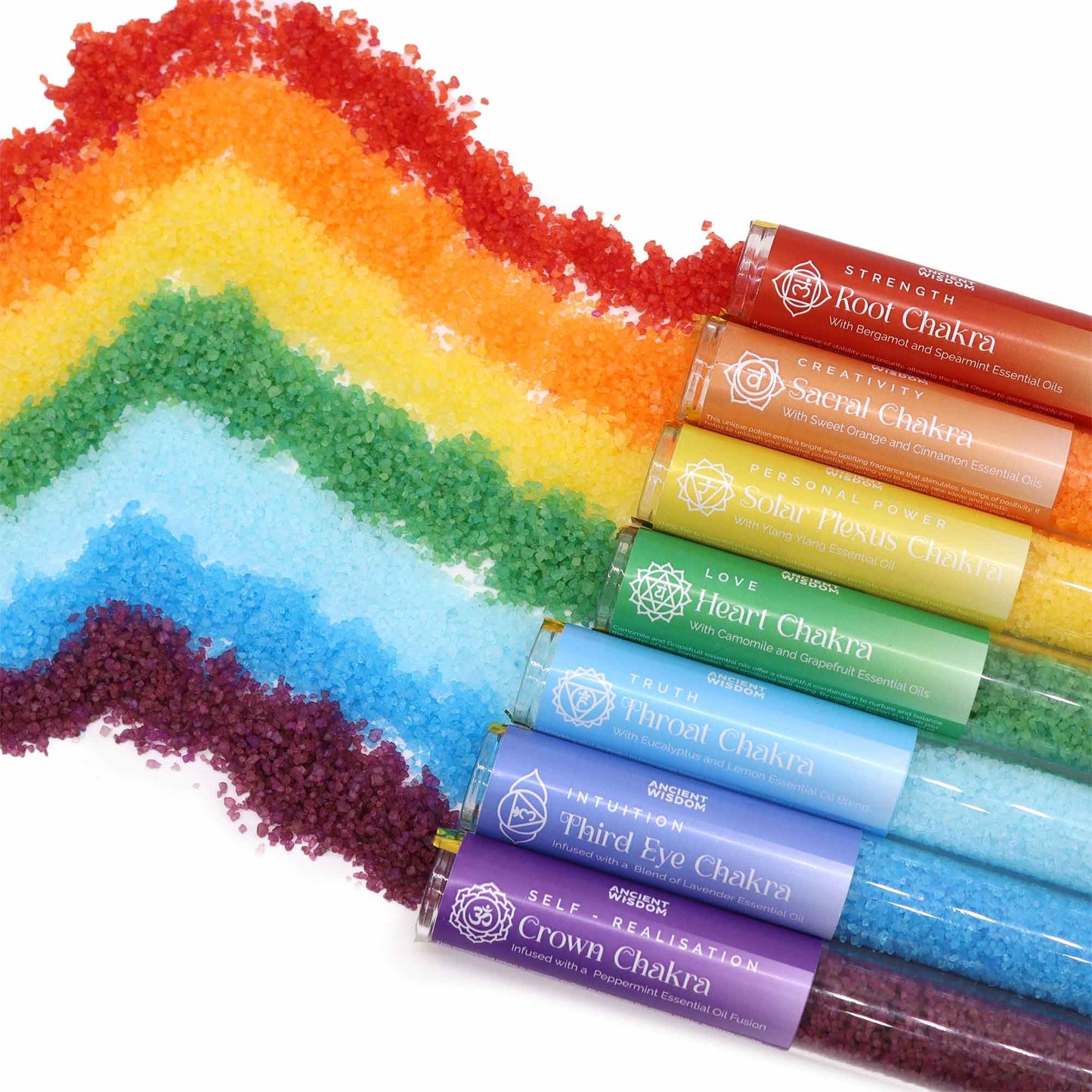 Rainbow row of 7 coloured chakra bath salt vials with essential oil droppers and crystals, arranged on white marble with candles and rose petals