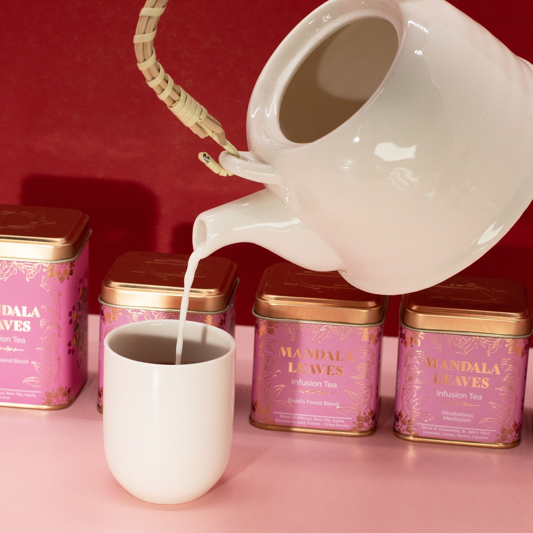 White teapot pouring milk into a white cup with 'Mandala Leaves' tea tins in the background.