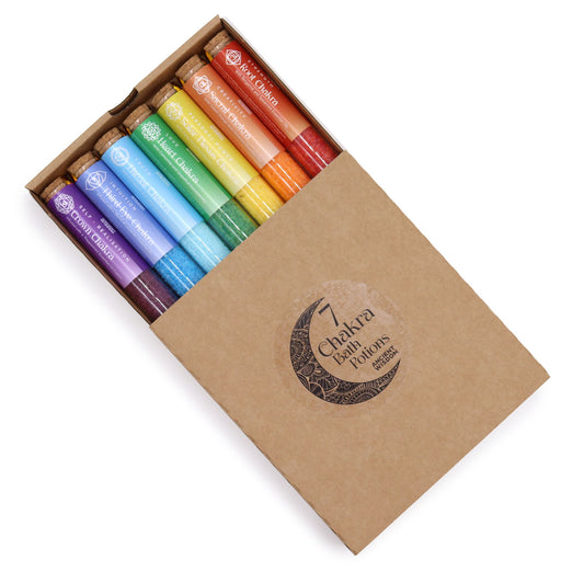 Rainbow row of 7 coloured chakra bath salt vials with essential oil droppers and crystals, arranged on white marble with candles and rose petals