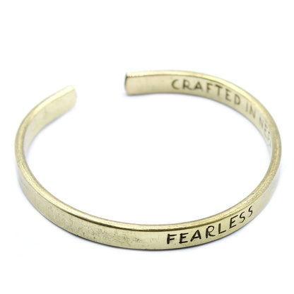 Golden brass mantra cuff engraved “FEARLESS” on wrist, Himalayan prayer flags and snow-capped peaks in soft background light