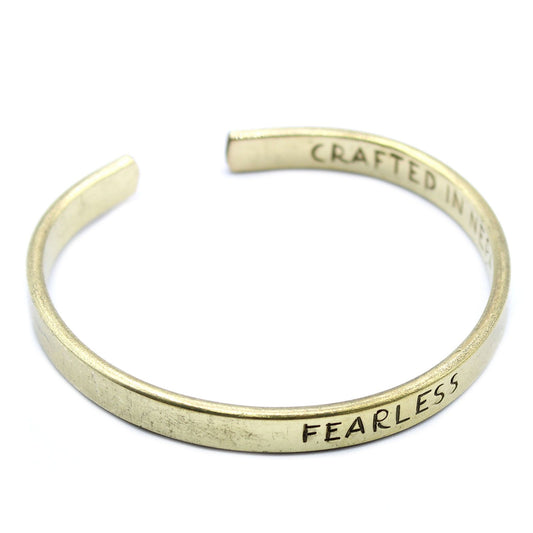 Golden brass mantra cuff engraved “FEARLESS” on wrist, Himalayan prayer flags and snow-capped peaks in soft background light