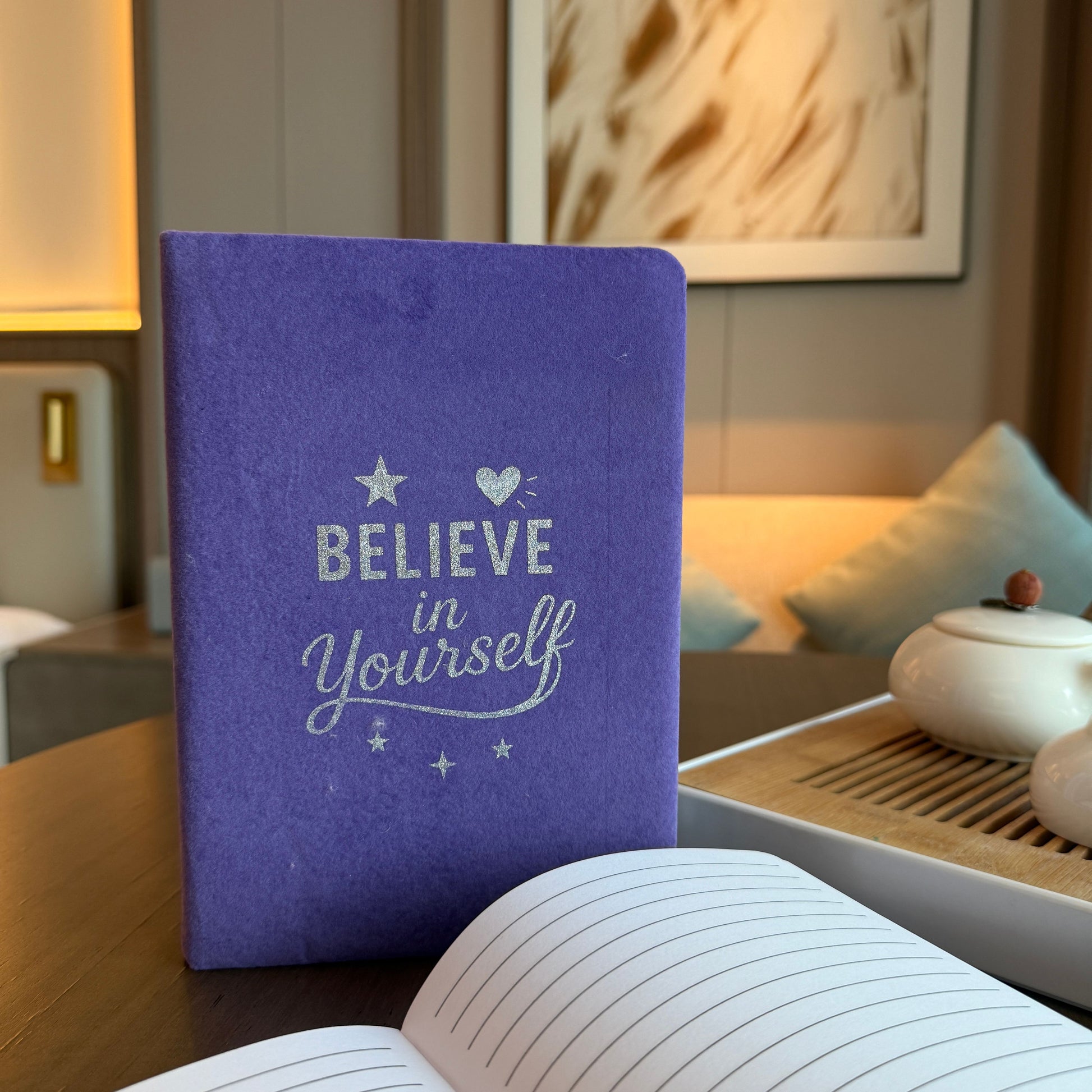 Purple notebook with 'Believe in Yourself' text on a wooden table in a cozy room.