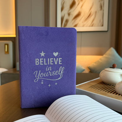 Purple notebook with 'Believe in Yourself' text on a wooden table in a cozy room.