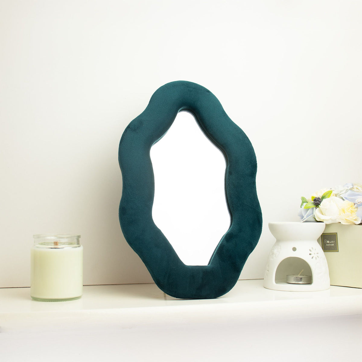 Soft squiggly oval pebble mirror in rich royal teal velvet frame with smooth curved edges, freestanding on white dressing table with soft natural light reflecting in glass