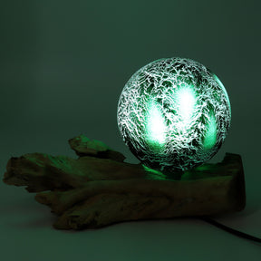 Emerald crackle glass sphere glowing atop unique twisted driftwood base on dark wooden side table with books and plants at night