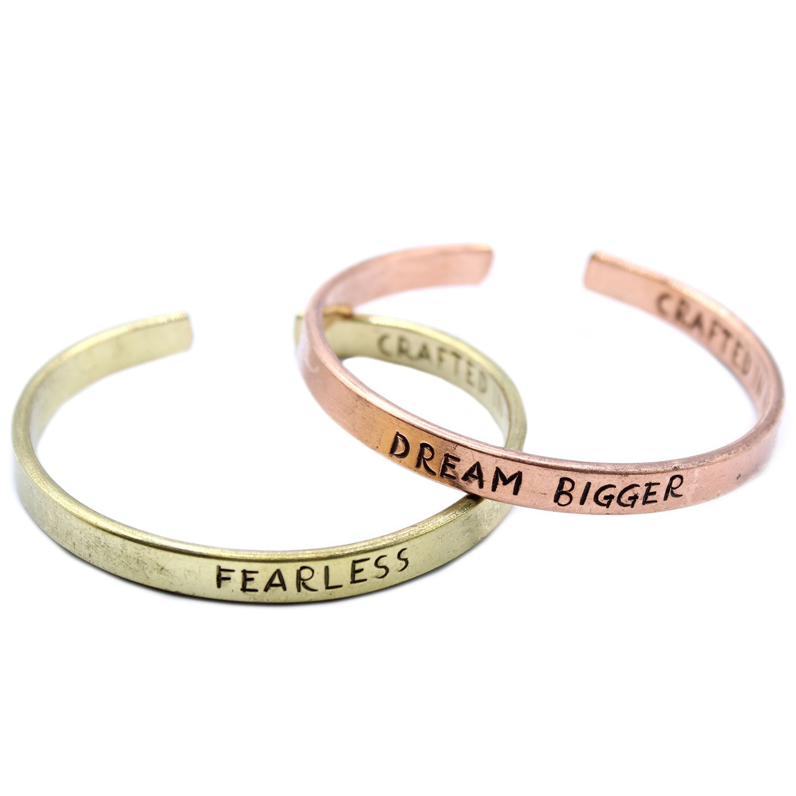 Open copper mantra cuff bracelet engraved “DREAM BIGGER” on wrist, surrounded by Himalayan prayer flags and mountains in soft sunrise light