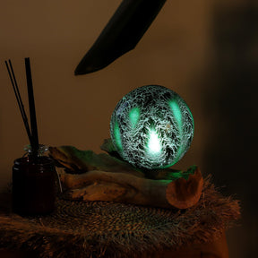 Emerald crackle glass sphere glowing atop unique twisted driftwood base on dark wooden side table with books and plants at night