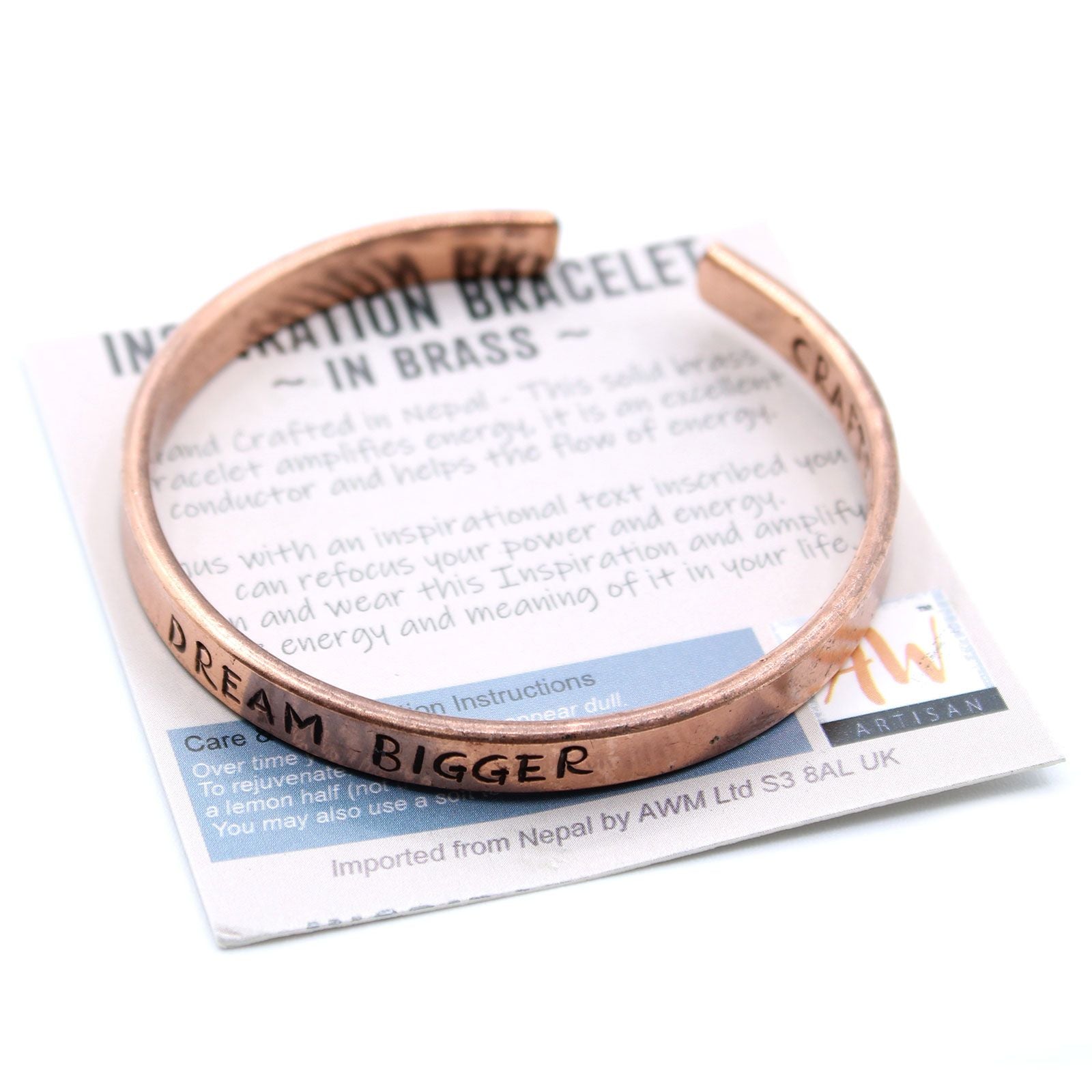 Open copper mantra cuff bracelet engraved “DREAM BIGGER” on wrist, surrounded by Himalayan prayer flags and mountains in soft sunrise light