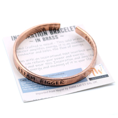 Open copper mantra cuff bracelet engraved “DREAM BIGGER” on wrist, surrounded by Himalayan prayer flags and mountains in soft sunrise light