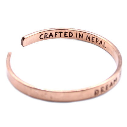 Open copper mantra cuff bracelet engraved “DREAM BIGGER” on wrist, surrounded by Himalayan prayer flags and mountains in soft sunrise light