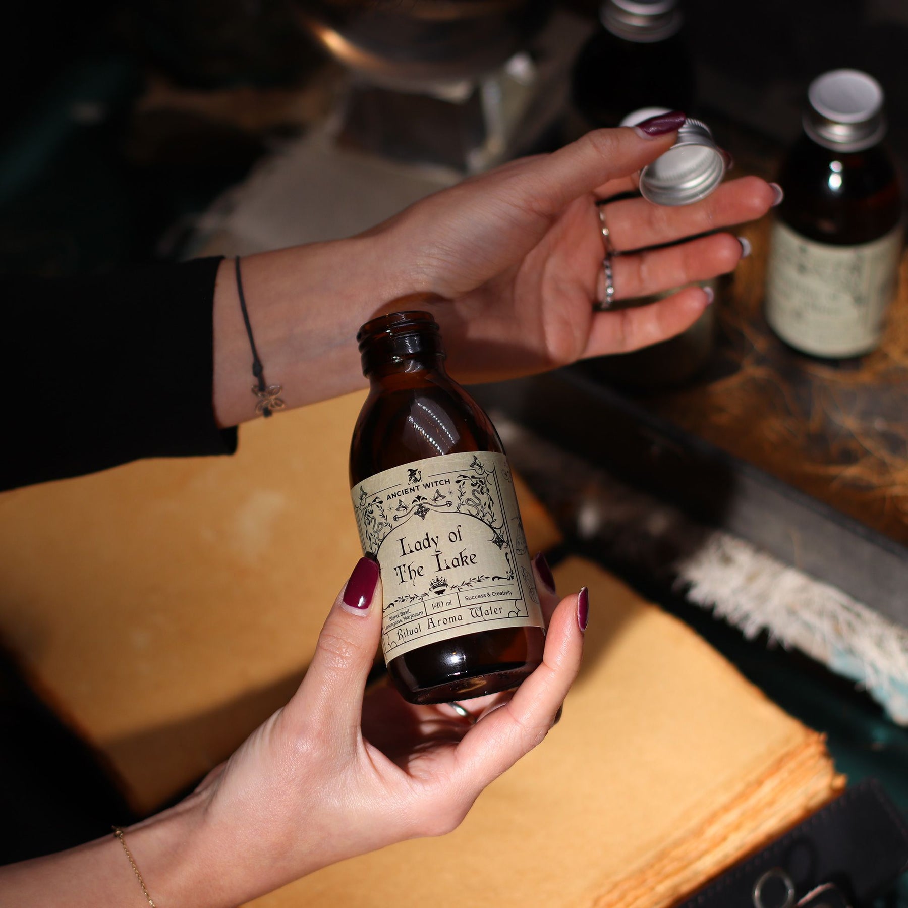 140ml amber glass bottle of Lady of the Lake ritual water with sword-in-lake label, surrounded by fresh basil, lemongrass, misty water and Excalibur sword