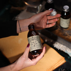 140ml amber glass bottle of Lady of the Lake ritual water with sword-in-lake label, surrounded by fresh basil, lemongrass, misty water and Excalibur sword