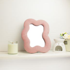 Soft squiggly square pebble mirror in warm rose blush velvet frame with gentle curved edges, freestanding on white dressing table with soft natural light reflecting in glass