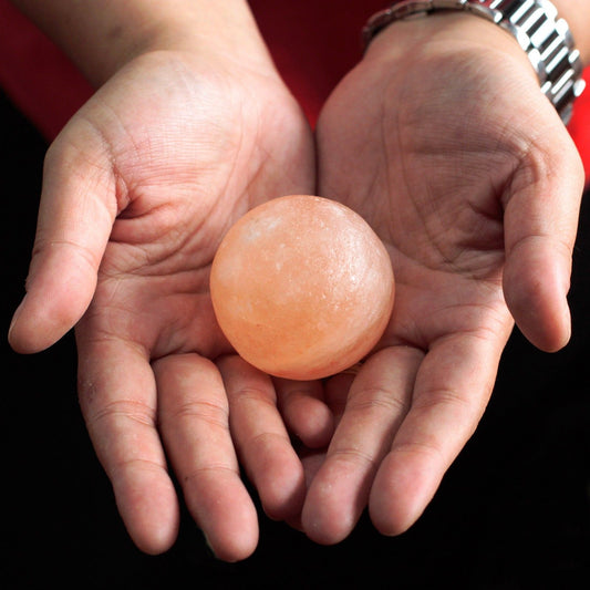 Two hands holding a Smooth ball-shaped Himalayan salt crystal deodorant stone against a black background