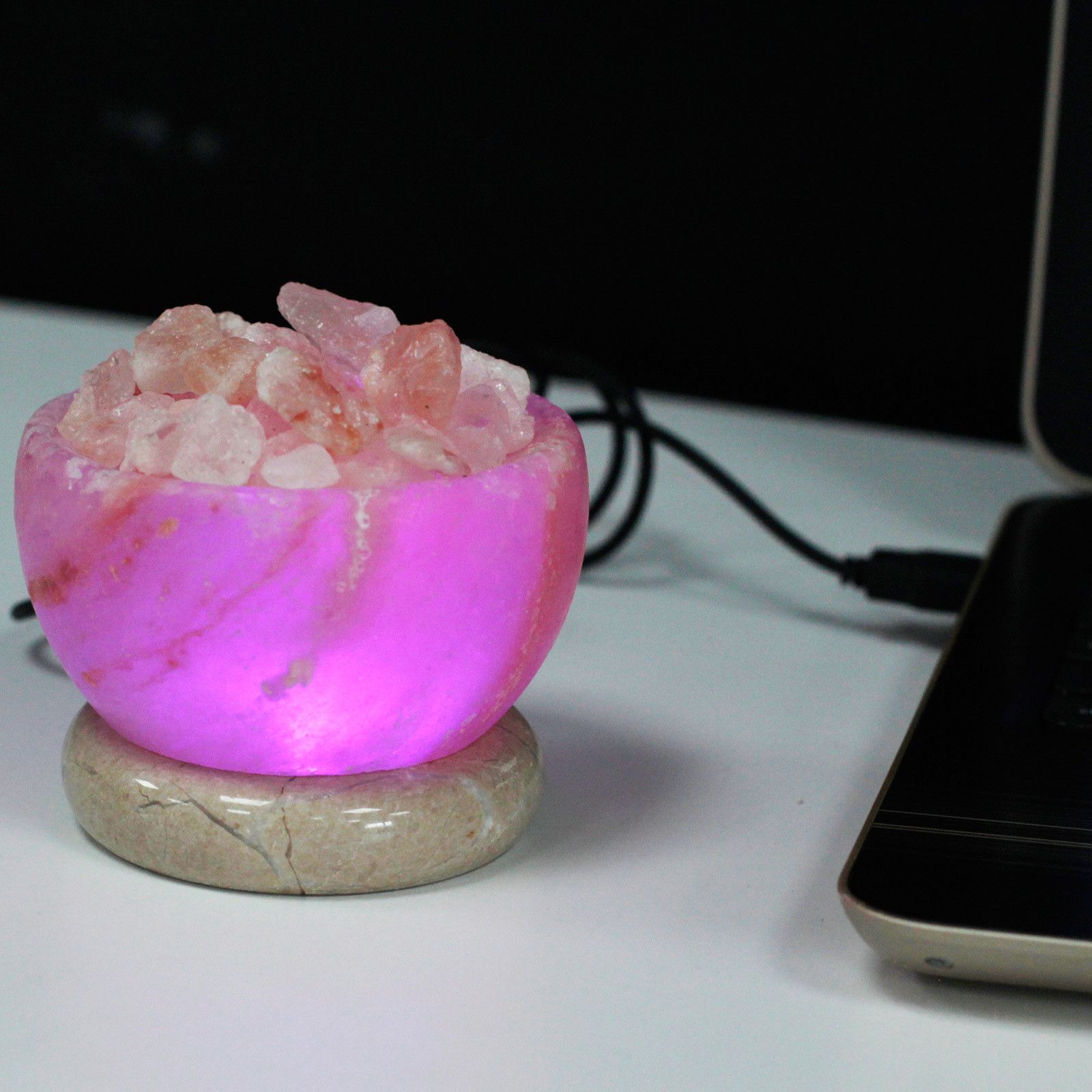 Himalayan salt fire bowl glowing with rotating rainbow colours, pink salt crystals lit from below, USB cable, on dark desk with laptop 
