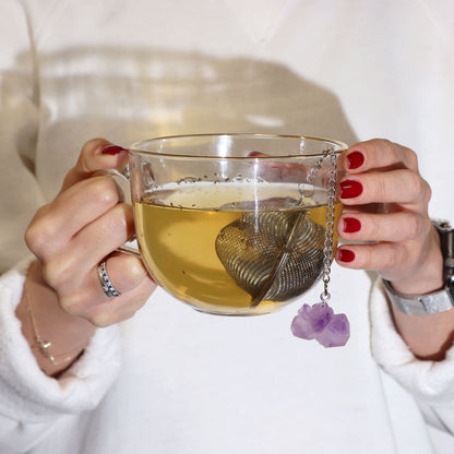 Person holding a glass mug of tea with a tea infuser and purple flower charm, wearing a white sweater.