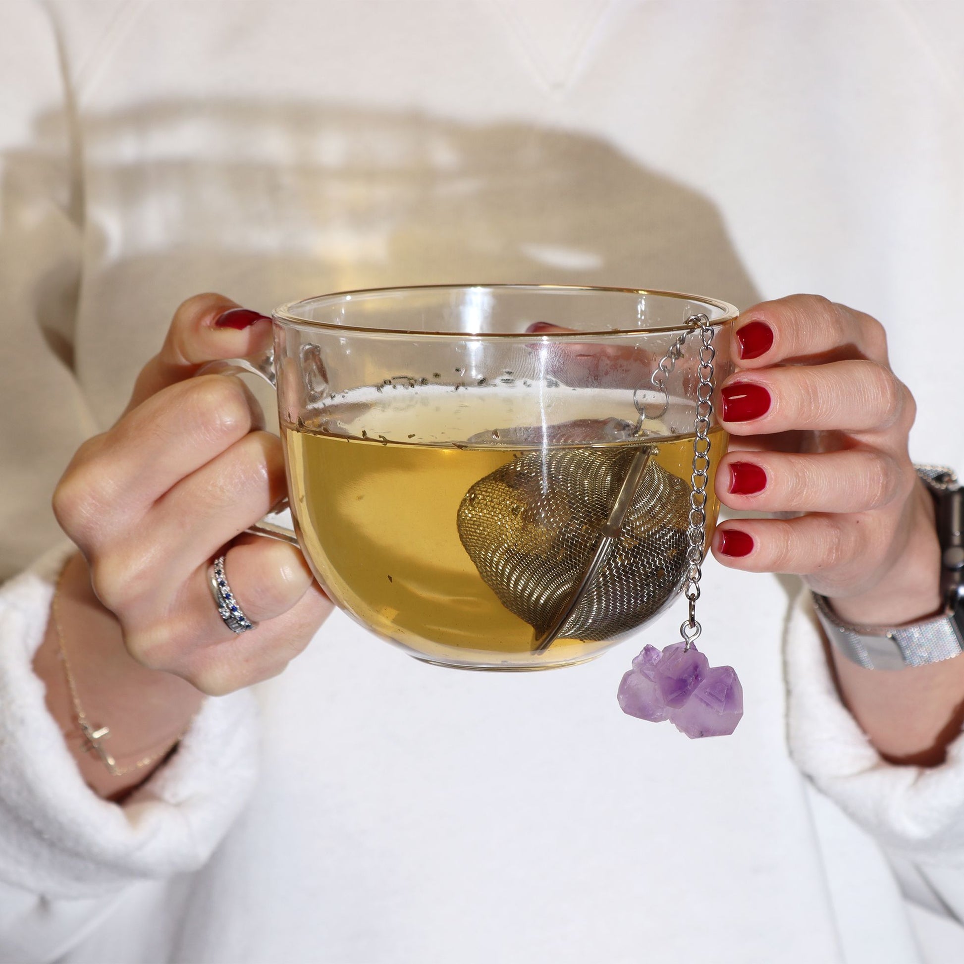 Person holding a glass mug of tea with a tea infuser and purple flower charm, wearing a white sweater.