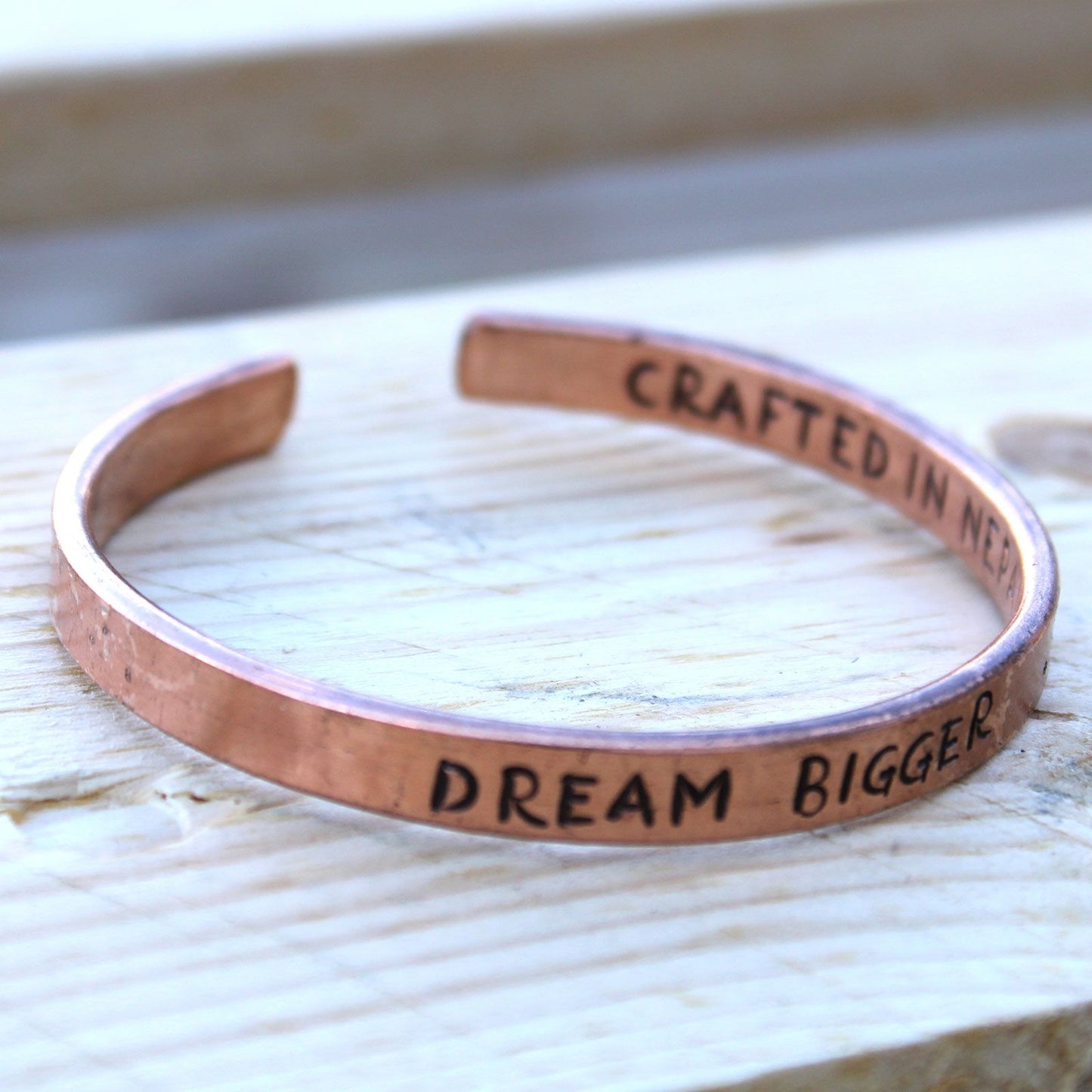 Open copper mantra cuff bracelet engraved “DREAM BIGGER” on wrist, surrounded by Himalayan prayer flags and mountains in soft sunrise light