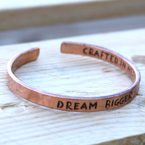 Open copper mantra cuff bracelet engraved “DREAM BIGGER” on wrist, surrounded by Himalayan prayer flags and mountains in soft sunrise light