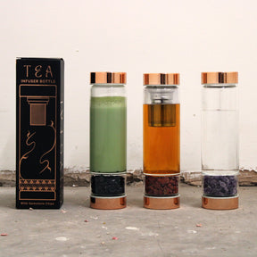 Three glass tea infuser bottles with different colored liquids and a packaging box on a textured surface.
