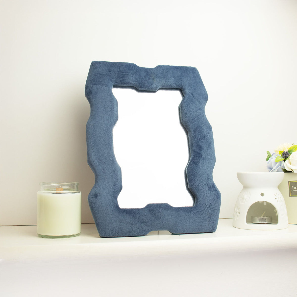 Soft squiggly mirror in rich royal blue velvet chunky frame with wavy sculpted edges, freestanding on white dressing table with soft natural light reflecting in glass