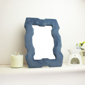 Soft squiggly mirror in rich royal blue velvet chunky frame with wavy sculpted edges, freestanding on white dressing table with soft natural light reflecting in glass
