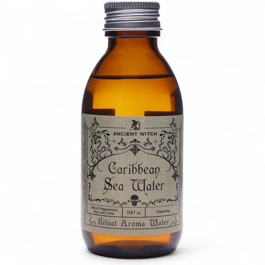 140ml amber bottle of Caribbean Sea Ritual Water with pirate-map label, surrounded by fresh limes, bay leaves, peppermint sprigs and ocean treasures