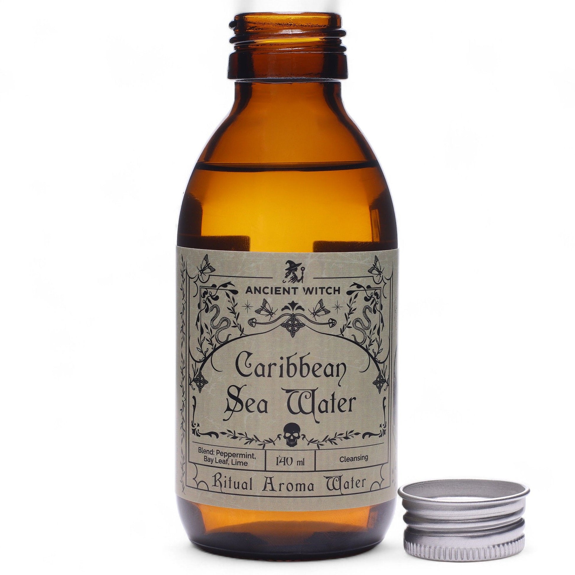 140ml amber bottle of Caribbean Sea Ritual Water with pirate-map label, surrounded by fresh limes, bay leaves, peppermint sprigs and ocean treasures