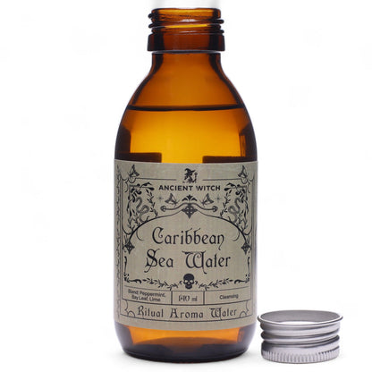 140ml amber bottle of Caribbean Sea Ritual Water with pirate-map label, surrounded by fresh limes, bay leaves, peppermint sprigs and ocean treasures