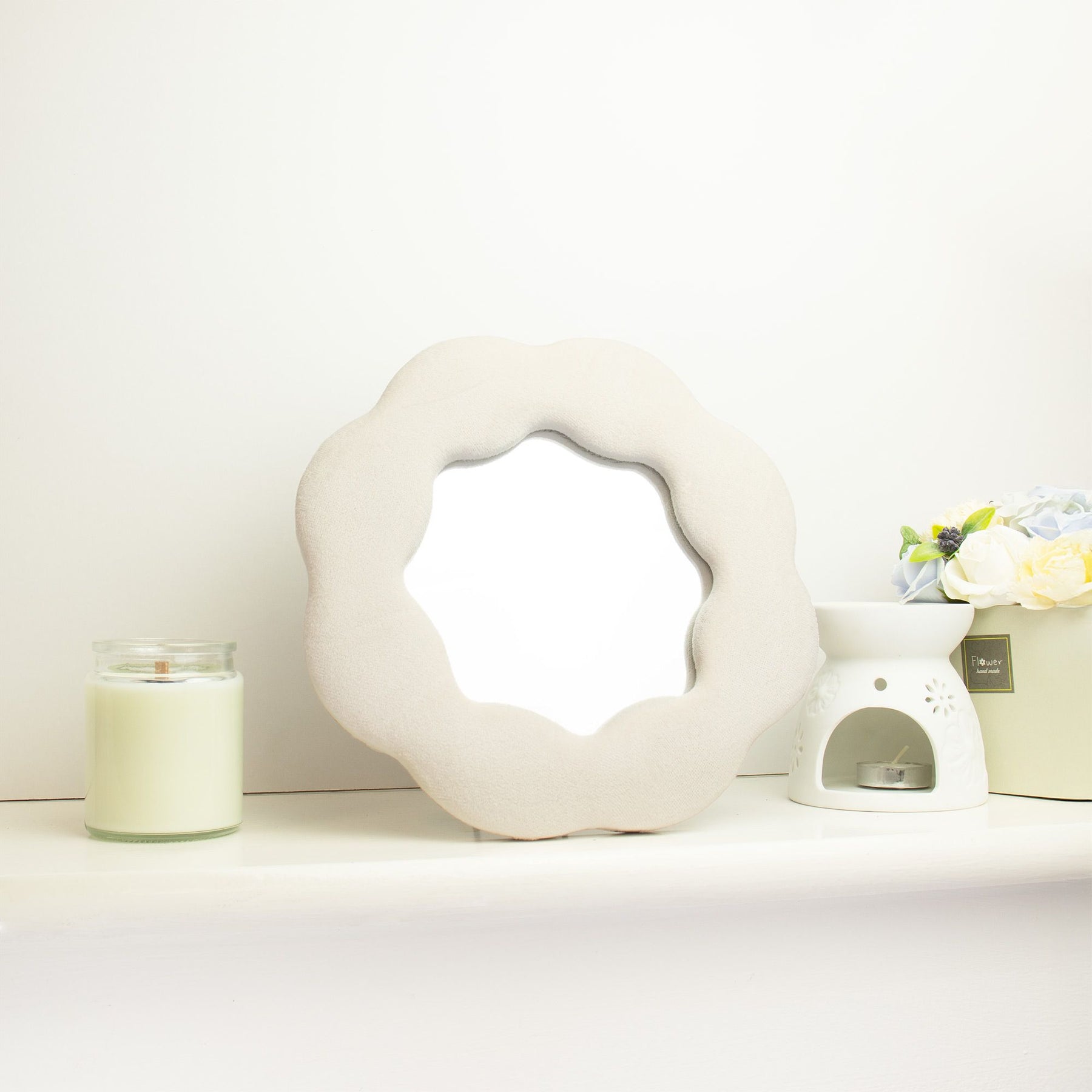 Soft squiggly mirror in neutral stone-toned velvet frame shaped like a gentle flower silhouette with wavy edges, freestanding on white shelf with soft natural light reflecting in glass