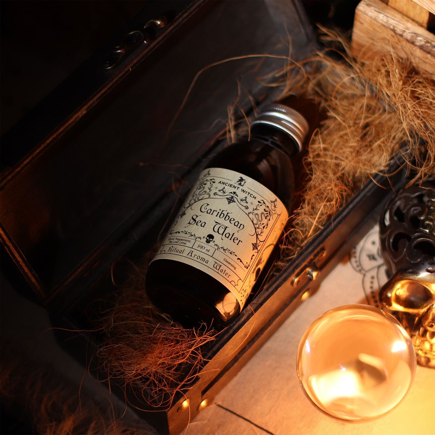 140ml amber bottle of Caribbean Sea Ritual Water with pirate-map label, surrounded by fresh limes, bay leaves, peppermint sprigs and ocean treasures
