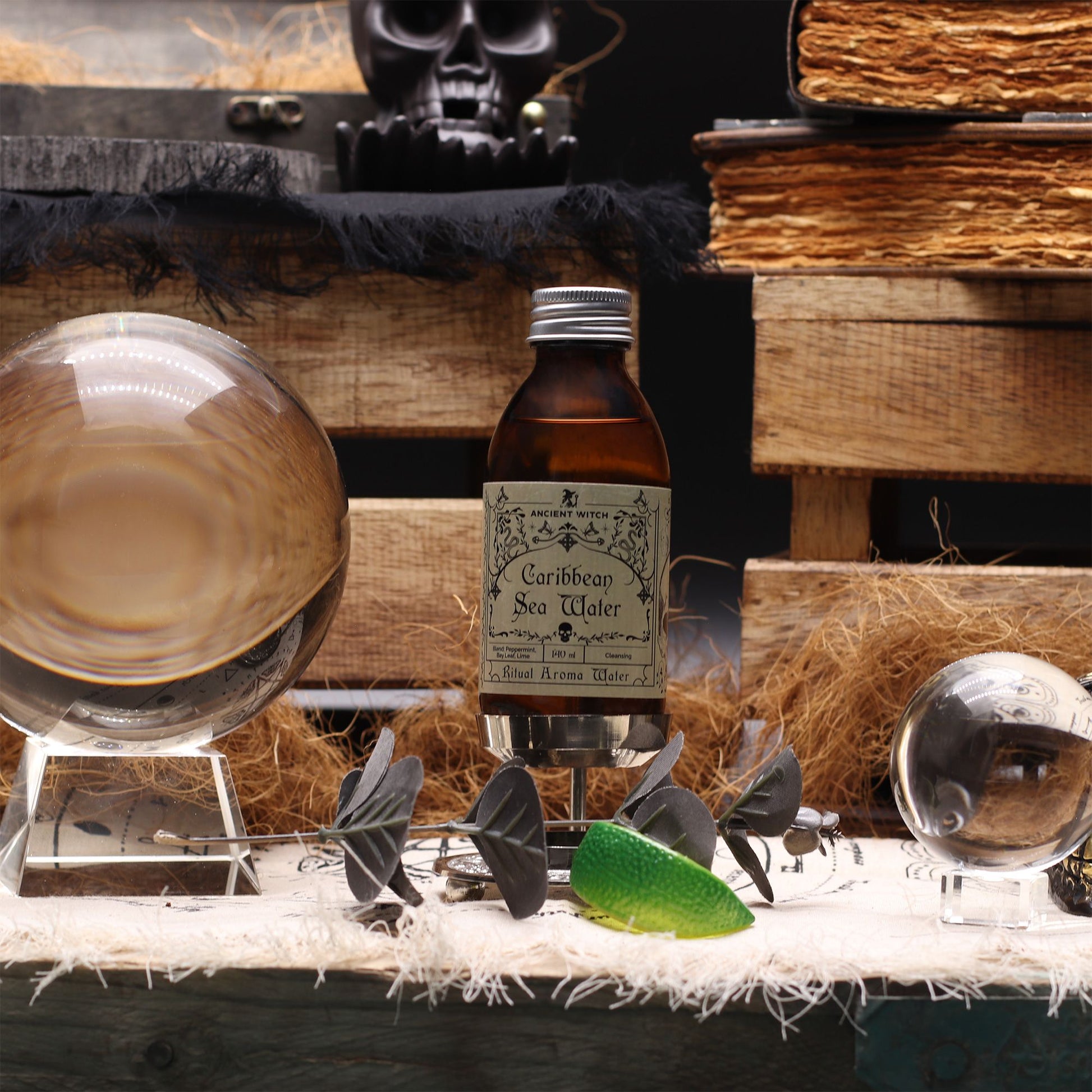140ml amber bottle of Caribbean Sea Ritual Water with pirate-map label, surrounded by fresh limes, bay leaves, peppermint sprigs and ocean treasures