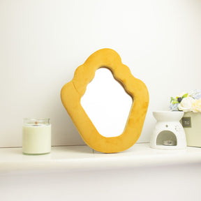 Soft squiggly mirror in vibrant marigold velvet frame with gentle wavy edges, freestanding on white dressing table with soft natural light reflecting in glass