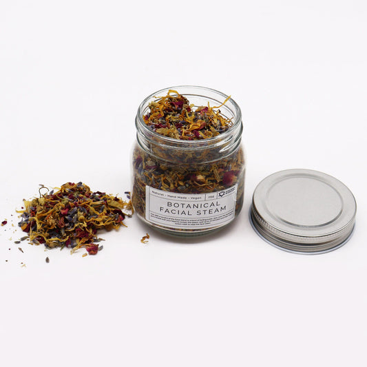 250g jar of dried lavender buds, rose petals, chamomile & calendula flowers spilling onto white marble with steaming bowl in soft spa setting